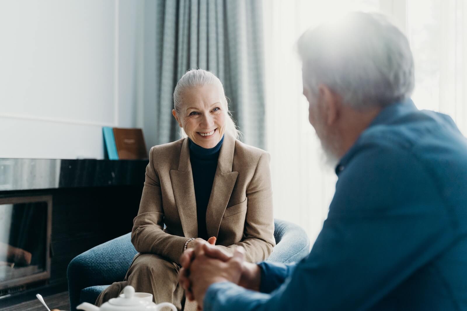 An older adult woman in business clothing smiles while having a discussion with another person, sitting in a casual office.