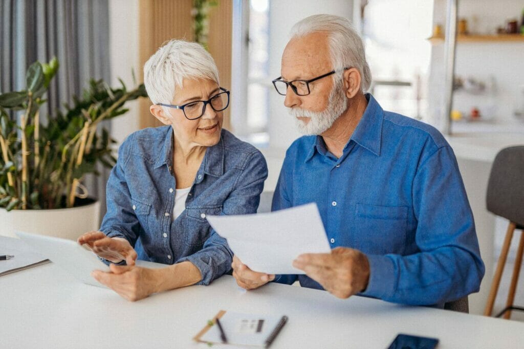 An older adult couple sits at a table while looking over and discussing paperwork.