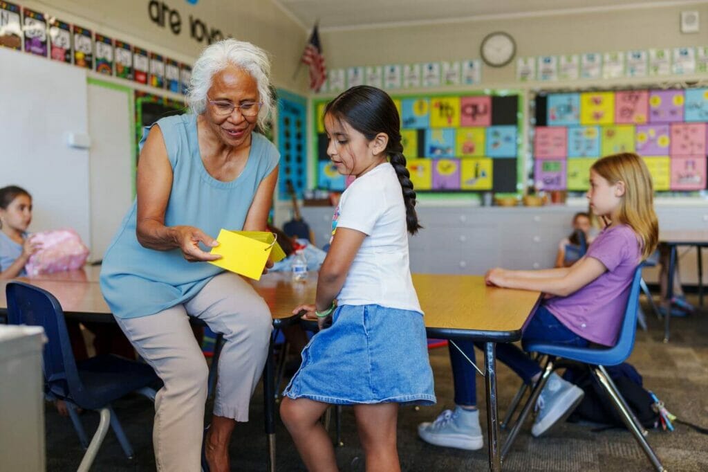 In a school classroom, an older adult woman sits on a table, helping a standing young girl with her schoolwork. There are other students in the background.