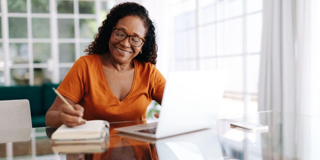 An older adult woman sits at a table in front of a laptop computer writing in a notebook.