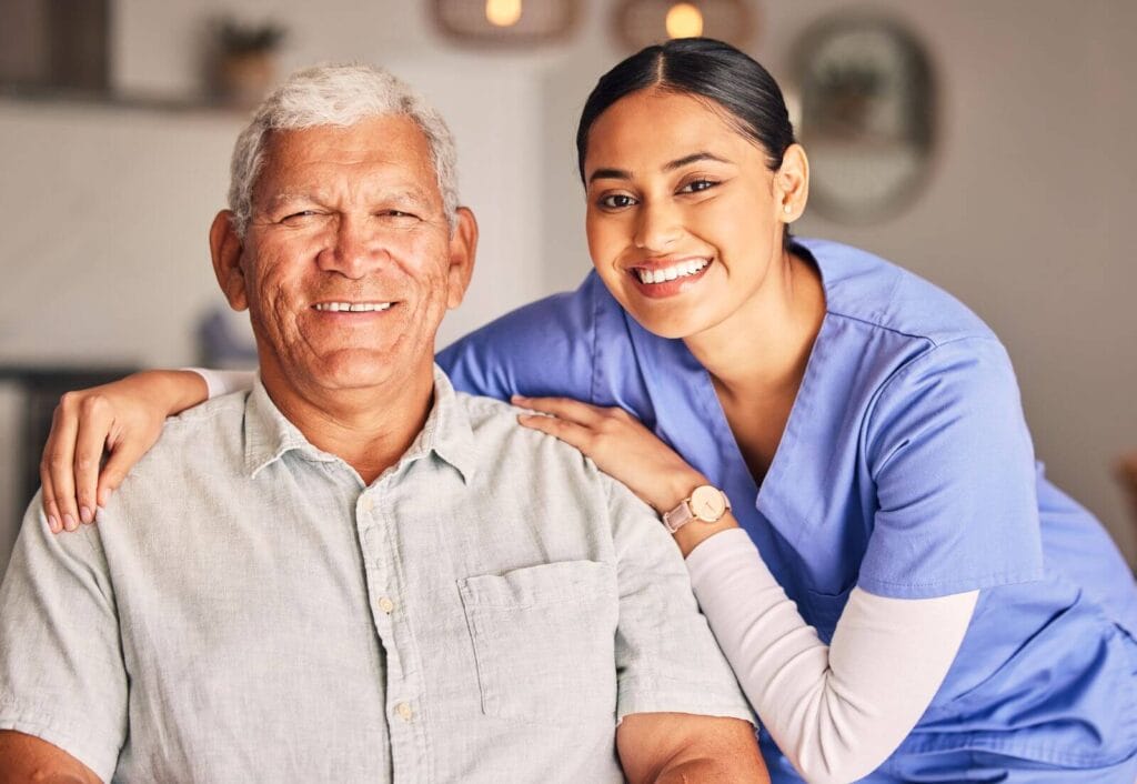 A woman wearing scrubs bends down to put her arms around a seated older adult man.