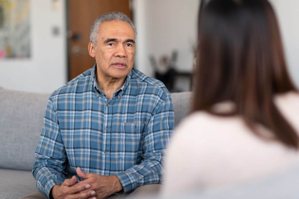 An older adult man sits on a couch, talking to a woman whose back is to the camera.
