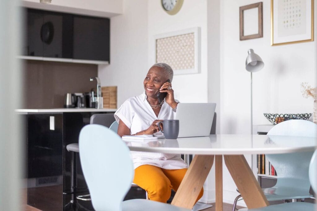 An older adult woman sits at a kitchen table in front of her laptop. She is talking on her cell phone and smiling.