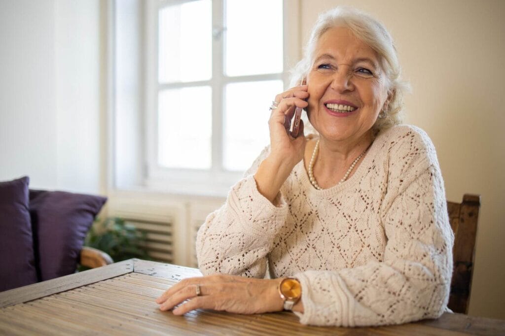 An older adult woman sits at at able, talking on her cell phone. She is smiling.