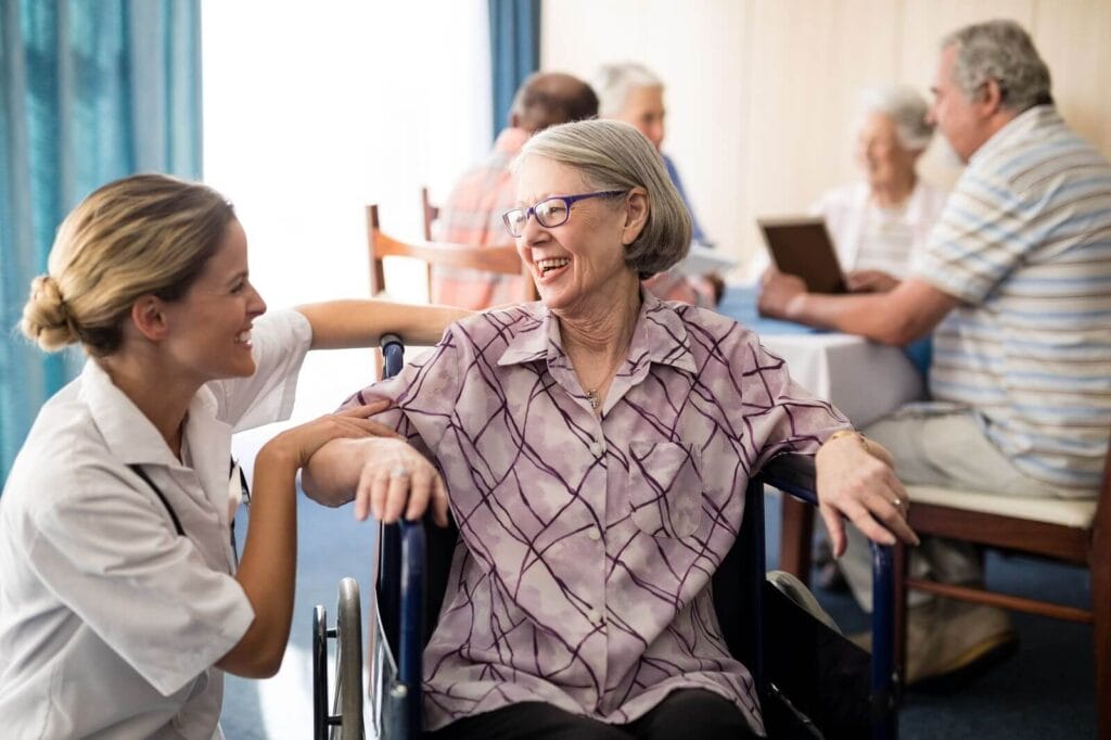 A woman kneels down next to and older adult woman sitting in a wheelchair. They smile at each other.