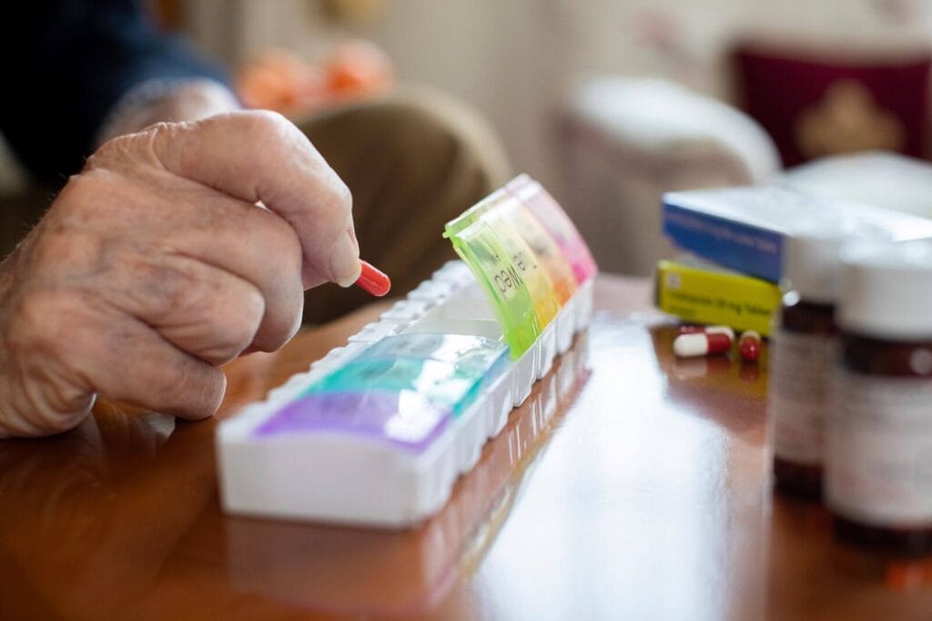 A closeup shows an older adult's hands placing pills into a weekly pillbox organizer.