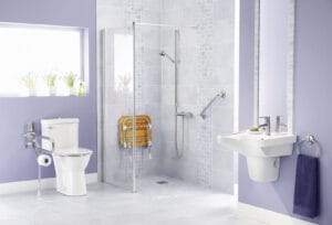 The image shows an empty bathroom with a sink, a walk-in shower with grab bars, a handheld showerhead, and a fold-down shower seat installed in the wall. There is also a toilet with a grab bar on one side.
