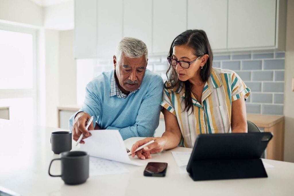 An older adult man and woman sit at a kitchen counter looking over paperwork together.