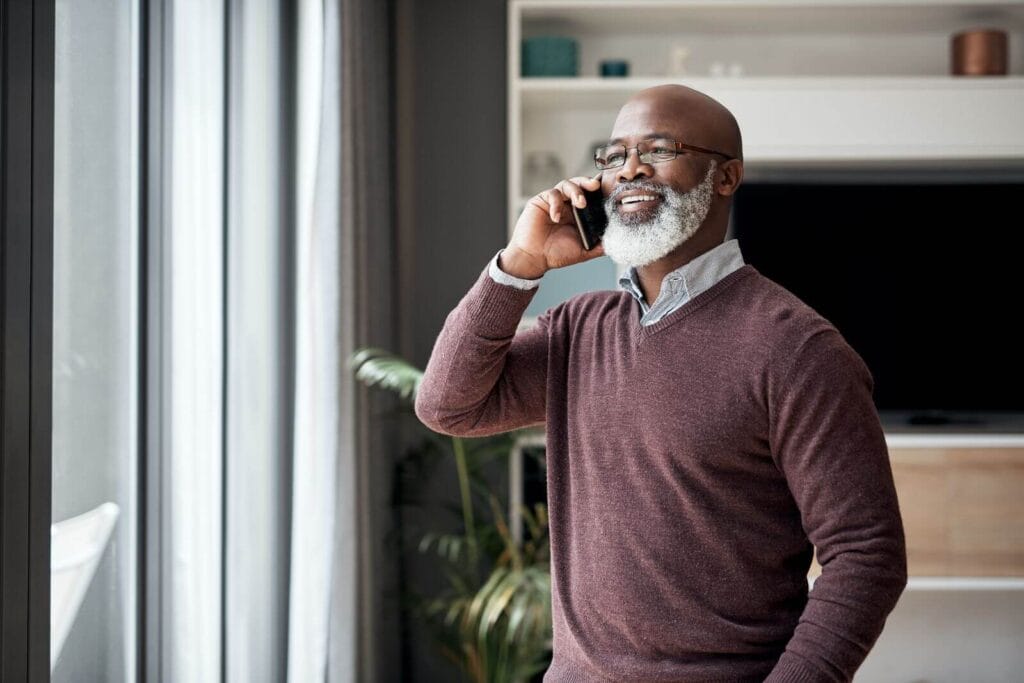 An older adult man looks out his window while talking on a cell phone.