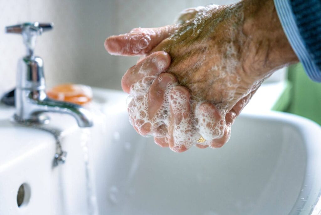 A close-up of an older adult man's hands as he washes them in the bathroom sink.