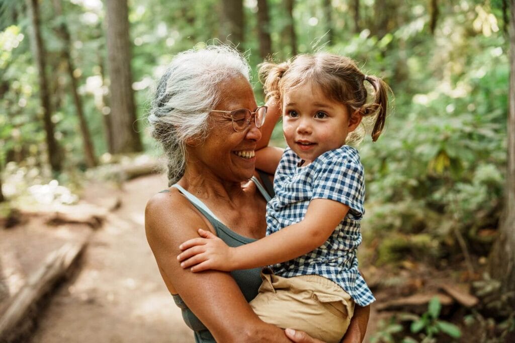 An older adult woman holds her young granddaughter in her arms. They are outside on a trail in the woods.