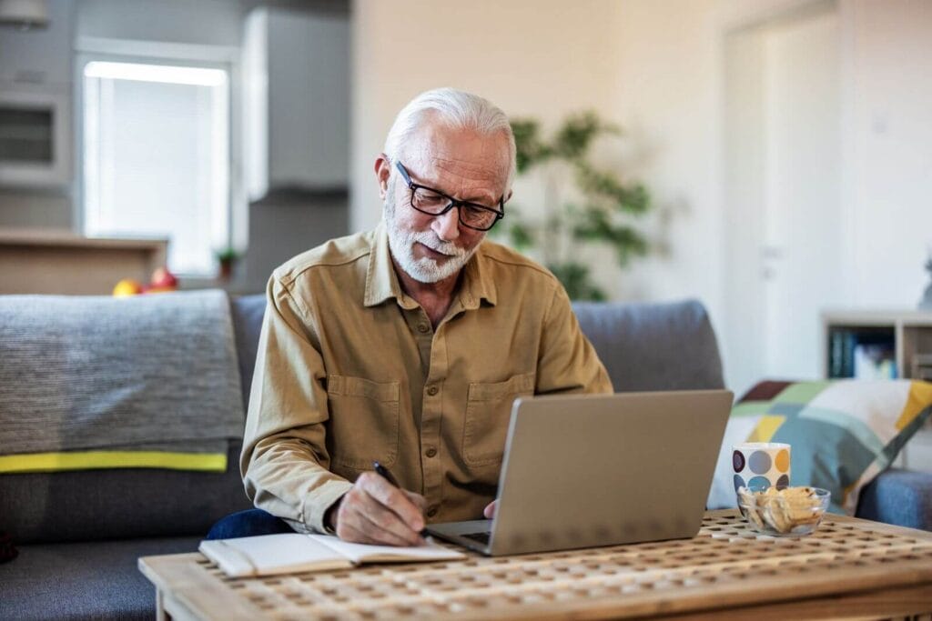An older adult man sits on a couch with a laptop and a notebook on a coffee table in front of him. He is taking notes in the notebook.