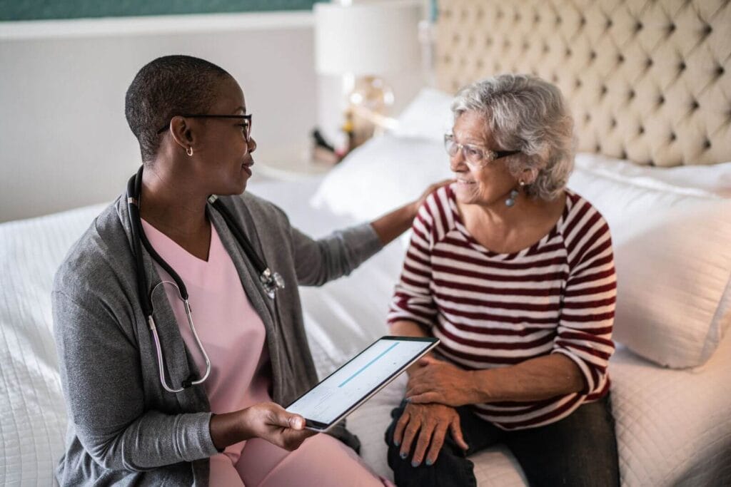 A woman health care professional wearing a stethoscope sits on a bed with an older adult woman. She is holding a tablet and putting her hand on the older woman's shoulder.