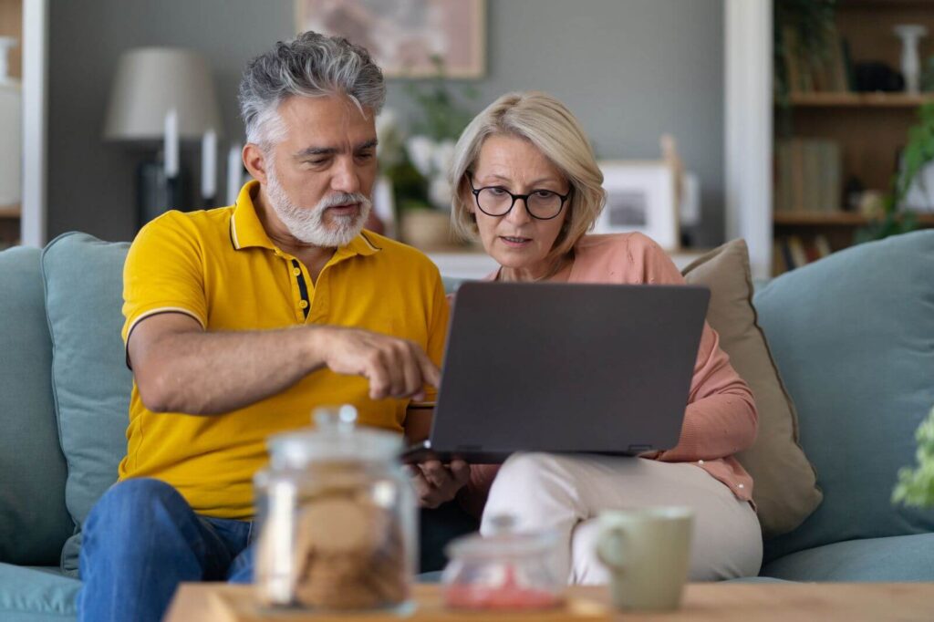 An older adult man and woman sit on a couch, looking at a laptop computer.