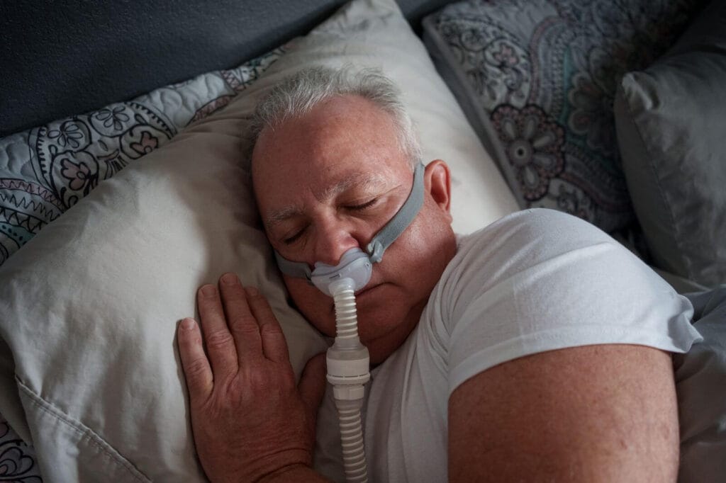 An older adult man sleeps in bed with a CPAP machine nasal mask on his nose.