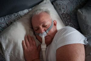 An older adult man sleeps in bed with a CPAP machine nasal mask on his nose.