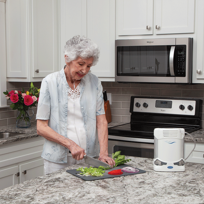 An older adult woman cuts vegetables in her kitchen. She is using the Caire FreeStyle Comfort portable oxygen concentrator.