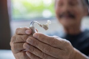 An older adult man holds a hearing aid up to the camera.