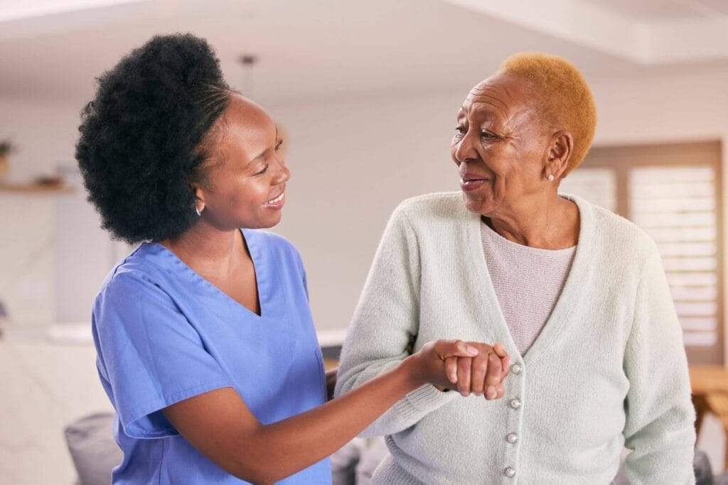 A female caregiver holds the arm of an older adult woman, helping her.