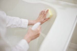 A woman's hands are shown holding a spray bottle and a sponge. She is cleaning the inner wall of a bathtub.