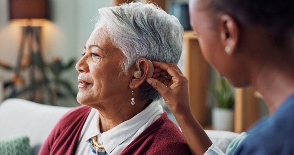 A female health care worker places a hearing aid in the ear of an older adult woman.