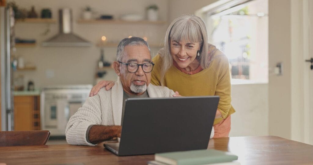 An older adult man sits at a table, typing on a laptop. An older adult woman is standing behind him, bending down and placing her hands on his shoulders.