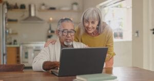 An older adult man sits at a table, typing on a laptop. An older adult woman is standing behind him, bending down and placing her hands on his shoulders.