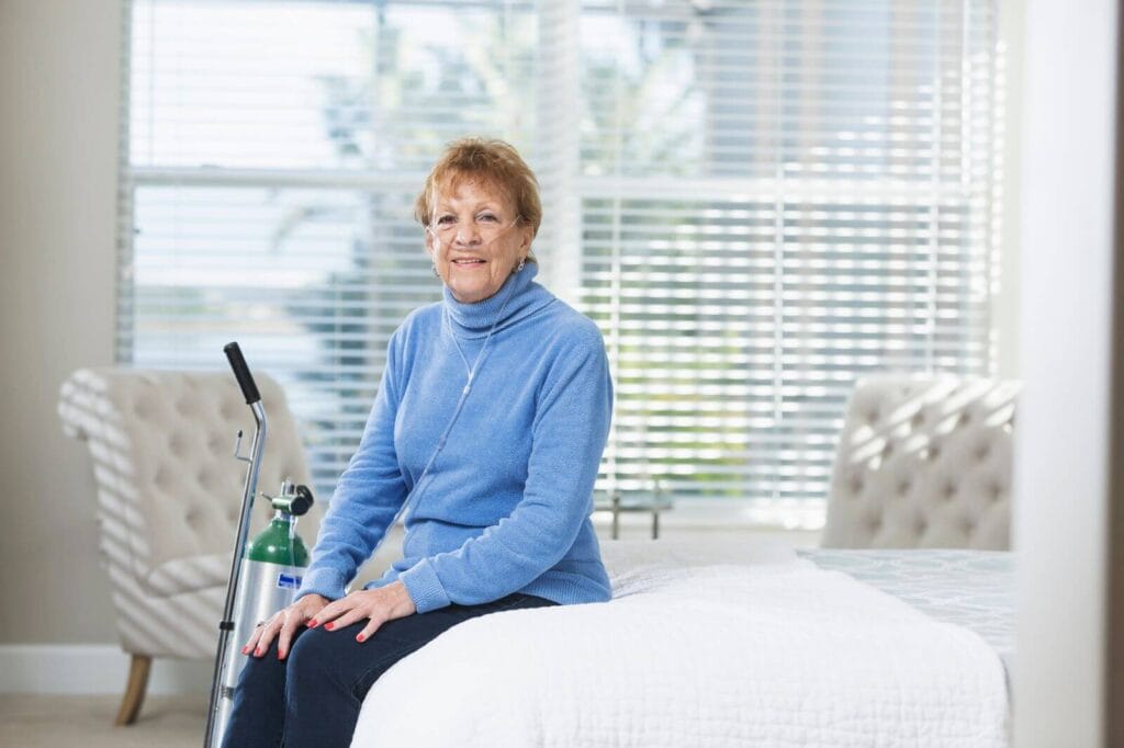 An older adult woman sits on the end of her bed. She has a nasal cannula in her nose and an oxygen tank beside her. She smiles.