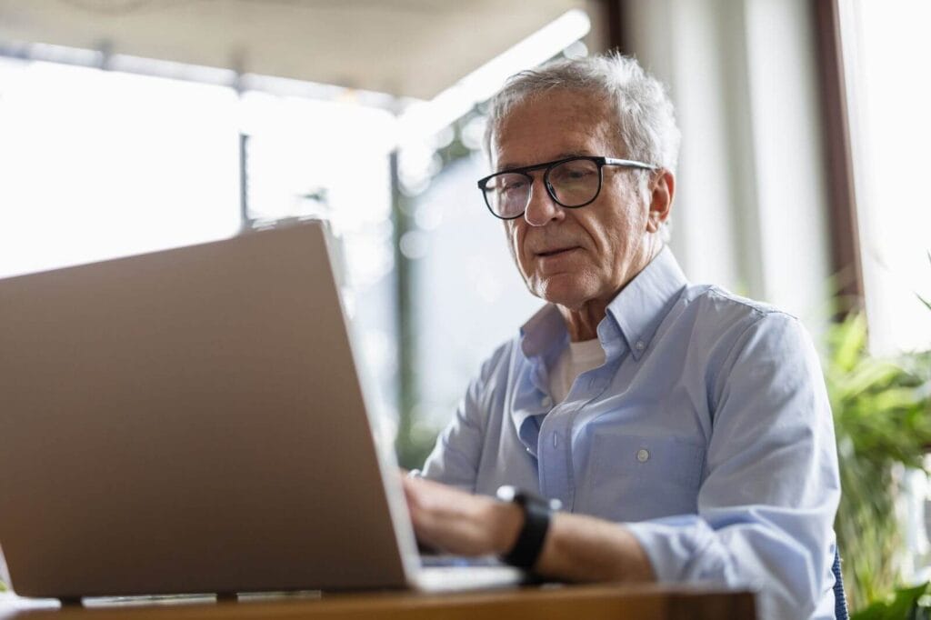An older adult man sits at a table using a laptop.