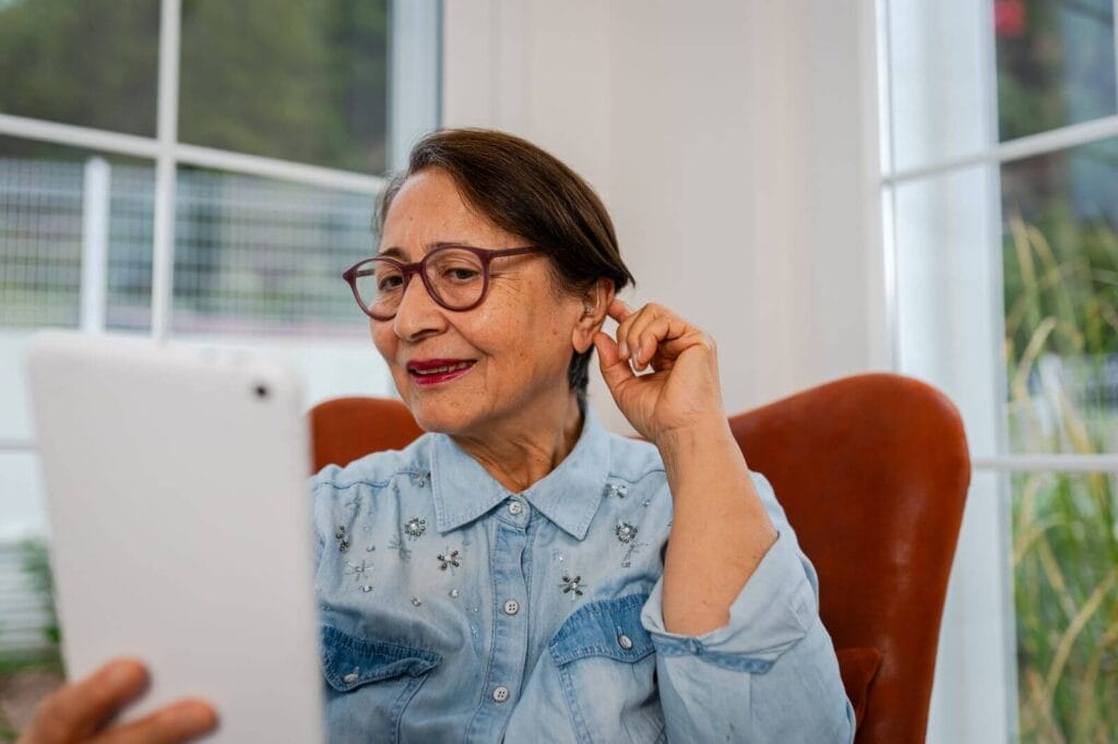 An older adult woman holds a hand up to her left ear, which has a hearing aid in it. She is holding a smartphone in front of her and looking at the screen.