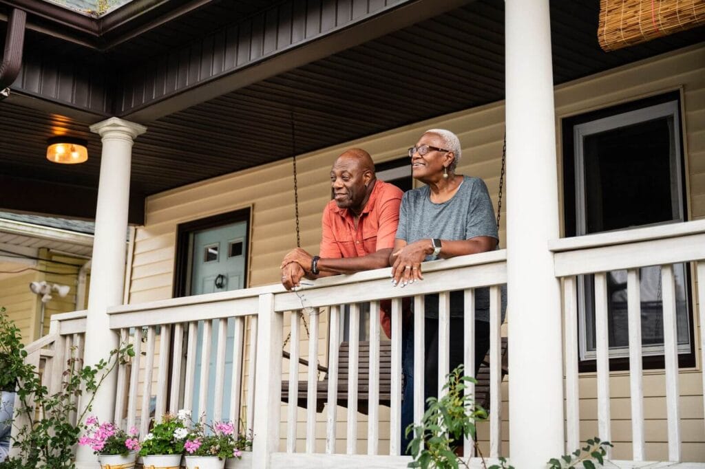An older adult man and woman stand on the front porch of their home, looking out into their front yard.