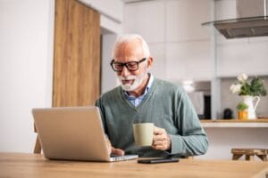 An older adult man sits at a kitchen table with a laptop in front of him. He is smiling and holding a coffee mug.