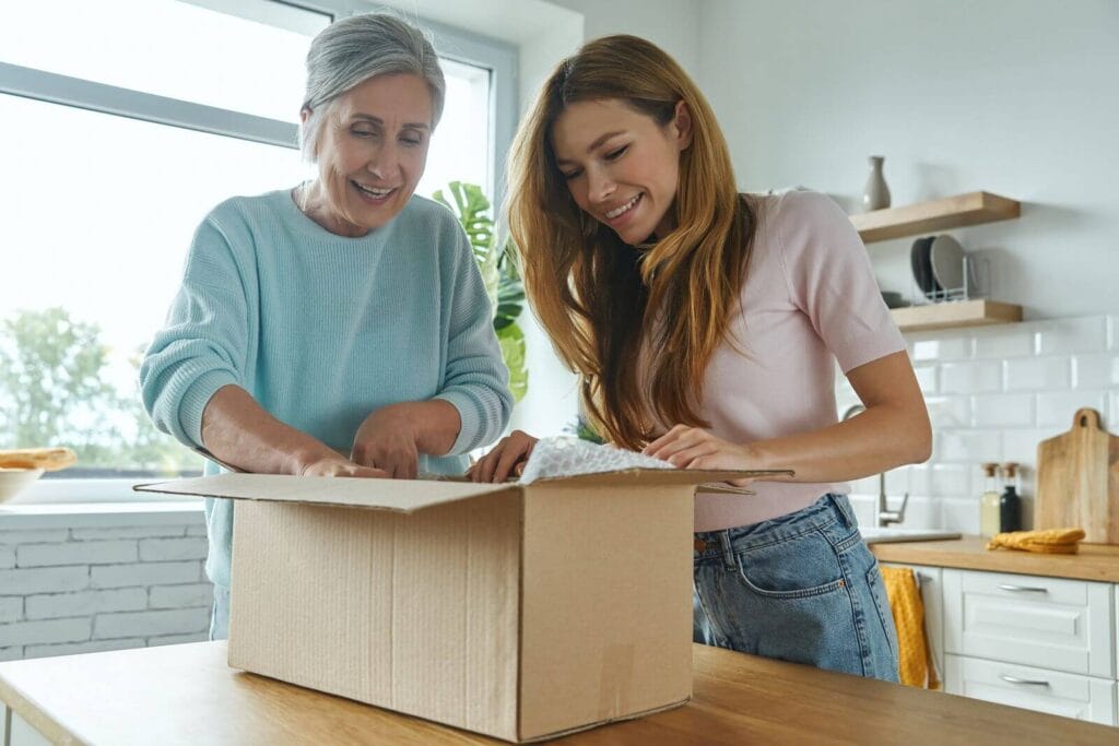 A woman and her older adult mother pack a box that is sitting on a kitchen island.