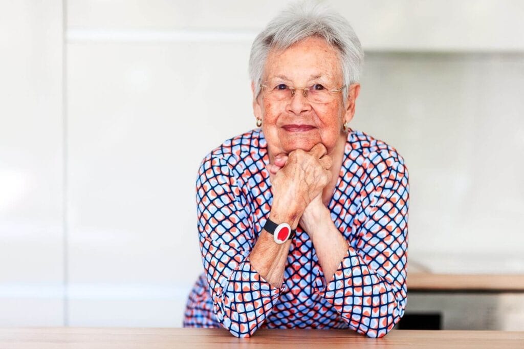 An older adult woman leans with her elbows on a counter. She is wearing a medical alert system pendant around her neck.