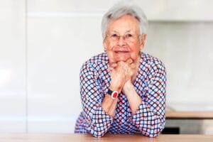 An older adult woman leans with her elbows on a counter. She is wearing a medical alert system pendant around her neck.