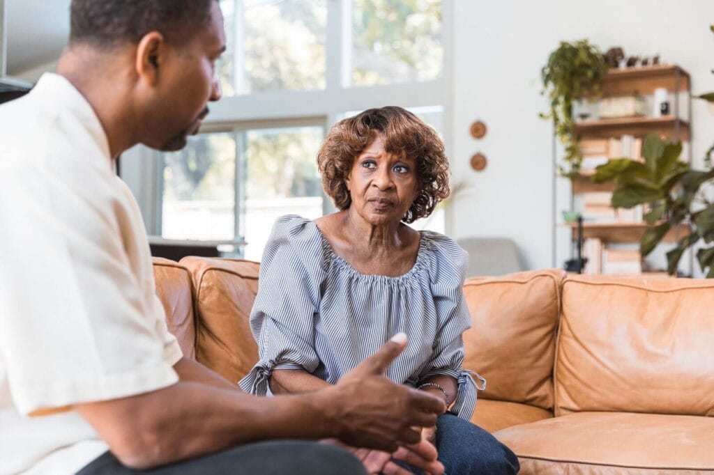 An older adult woman and her son sit on a couch, having a discussion.