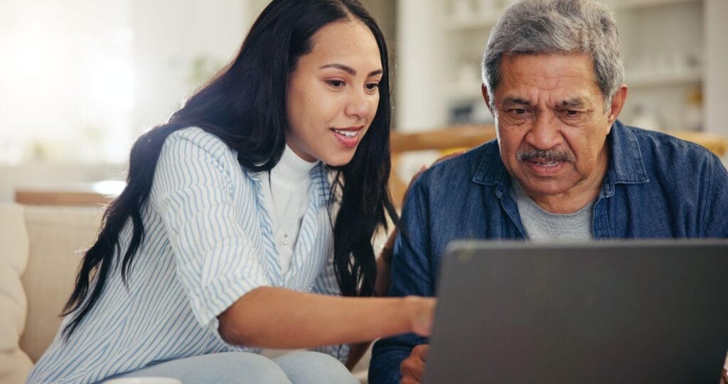 A woman sits on a couch with her older adult father. She is pointing to something on a laptop on a table in front of them.
