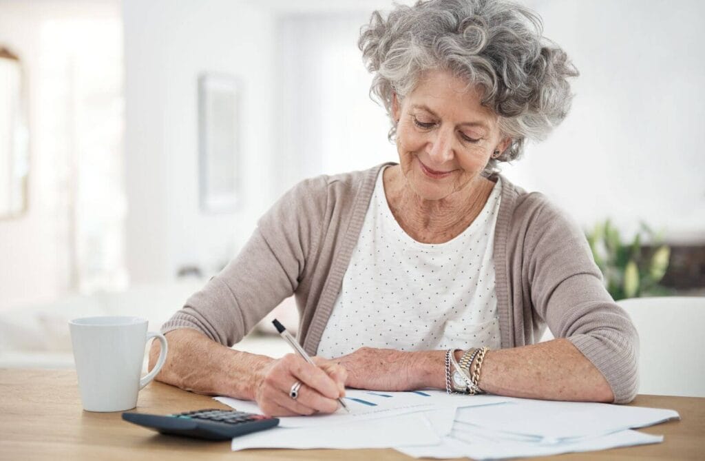 An older adult woman sits at a table, writing on some paperwork. There is a calculator and a coffee mug on the table.