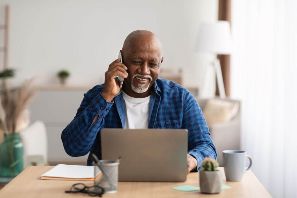An older adult man sits at a table, talking on his cell phone. There is a laptop, a coffee mug, a notebook, a pen holder, a small plant, and a pair of glasses on the table.