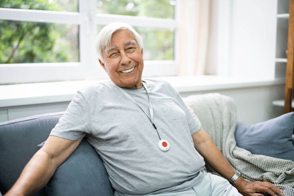 An older adult man sits on a couch, smiling at the camera. He is wearing a medical alert system pendant around his neck.