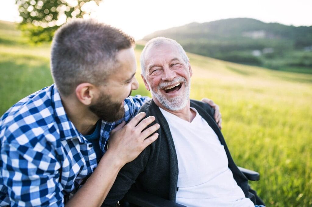 An older adult man sits in a wheelchair in a grassy field. His son is behind him, crouched down with his hands on his father's shoulders. They smile at each other.