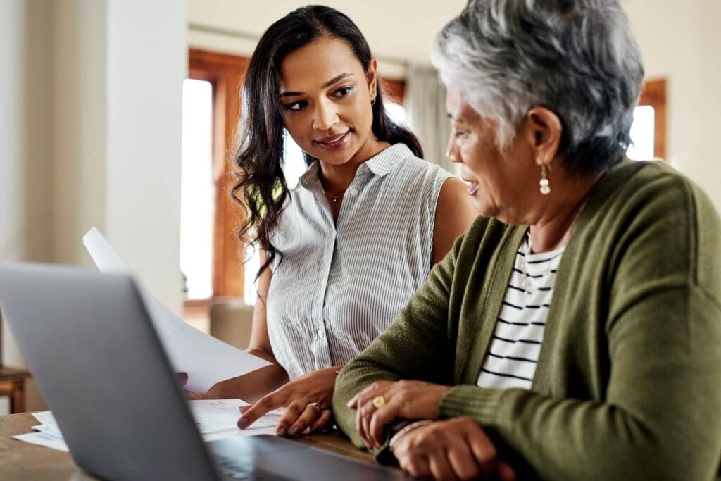 A woman sits at a desk or table with her older adult mother. They are looking at paperwork and have a laptop in front of them.
