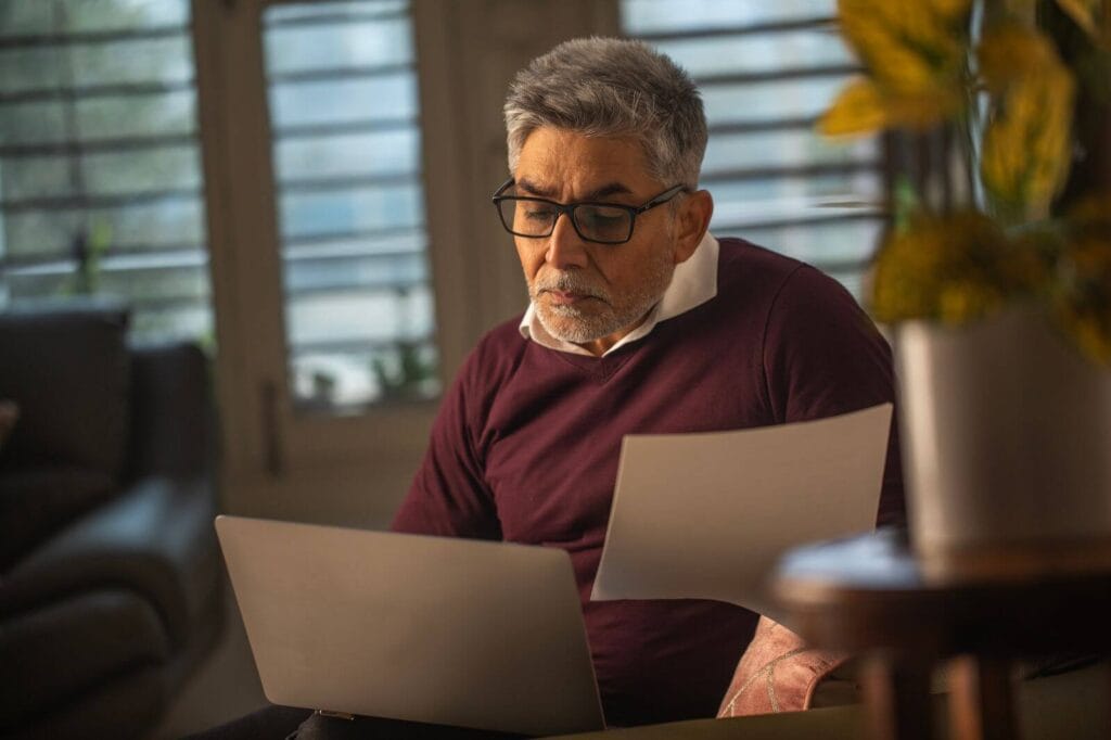 An older adult man sits, holding paperwork. He has a laptop on his lap.