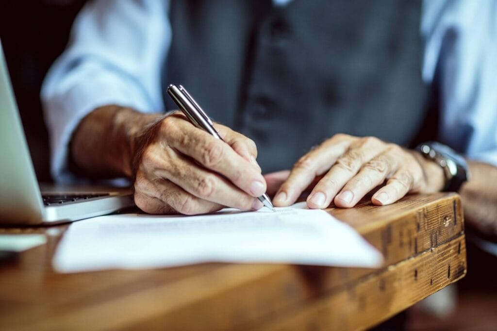 An older adult man's hands are shown in a close-up signing paperwork.