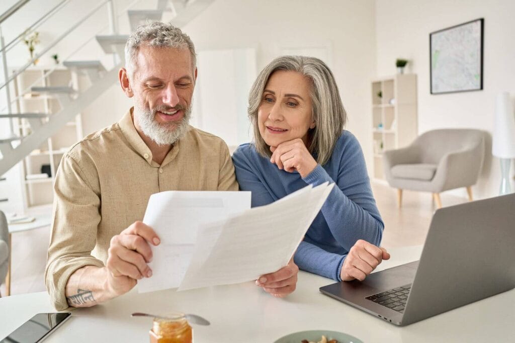A middle-aged man and woman sit at a table together looking at paperwork. There is a laptop in front of them.