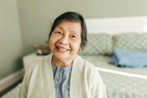 An older adult woman is standing in her bedroom with the bed behind her. She smiles.
