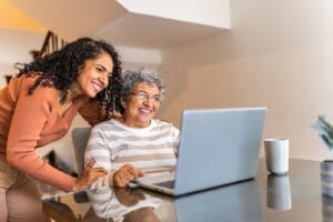 An older adult woman is sitting at a table with a laptop and a coffee mug in front of her. Her daughter is standing behind her, leaning to see the screen. They are smiling.