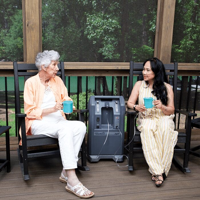 Tw women sit on a porch with coffee mugs. They are both using the NewLife Intensity oxygen concentrator simultaneously.