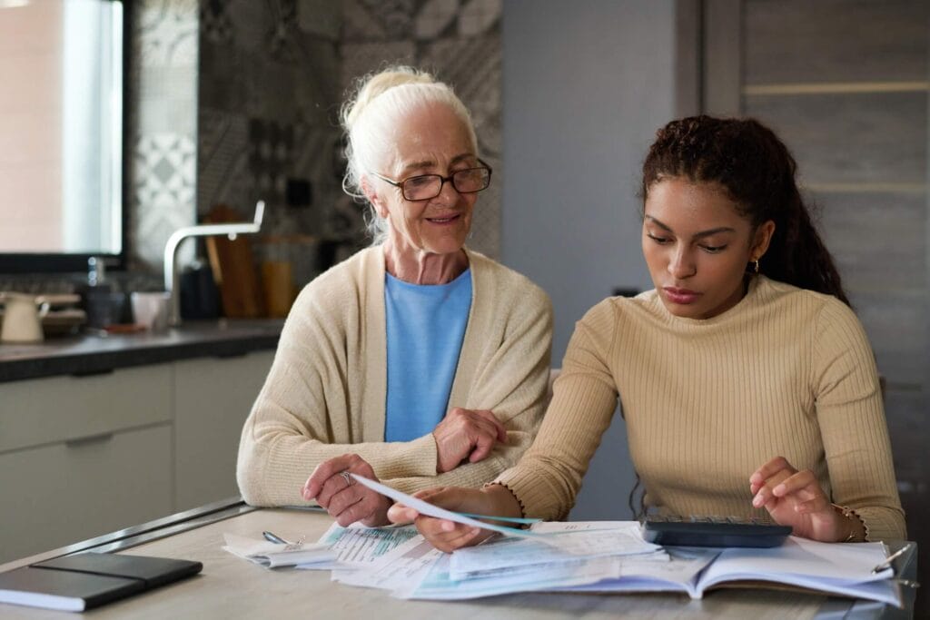 A woman sits at the kitchen table with her older adult mother. There is paperwork and a calculator on the table in front of them.