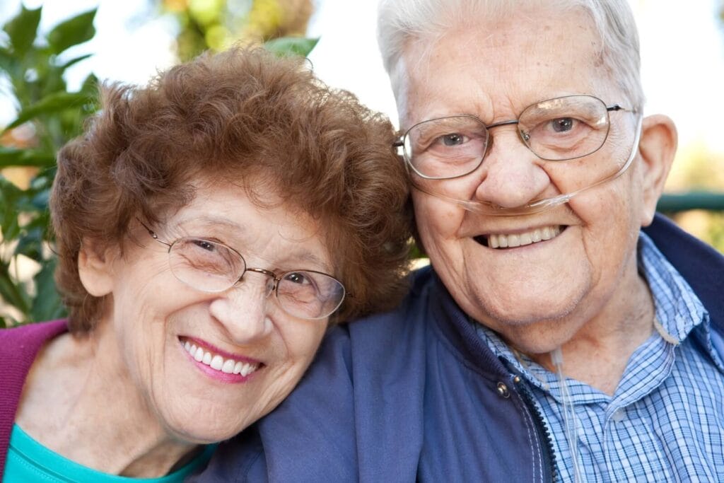 An older adult man and woman smile at the camera. The man is wearing a nasal cannula for supplemental oxygen.